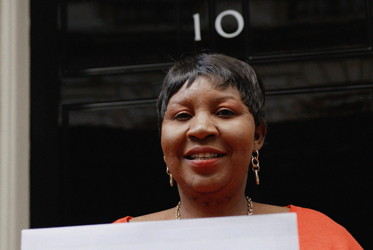 A woman stands smiling holding a piece of paper in front of Number 10 Downing Street. She is wearing an orange top and gold jewelry. Behind her is a black door with the number ten on it. 
