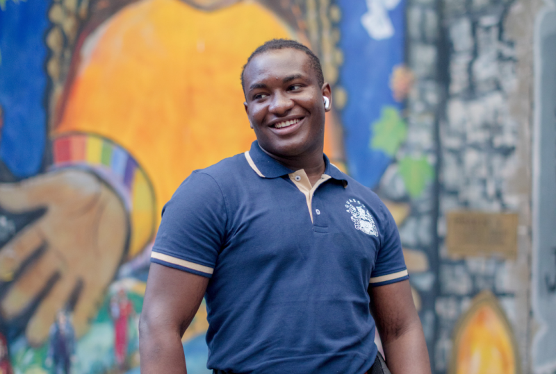 A young black man stands in the middle smiling. In the background is a colourful mural on a building wall.