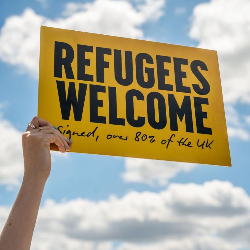 The image shows a yellow sign held up against a partly cloudy sky. The sign has bold, black text that reads: "REFUGEES WELCOME." Below this, in handwritten-style text, it says: "Signed, over 80% of the UK."